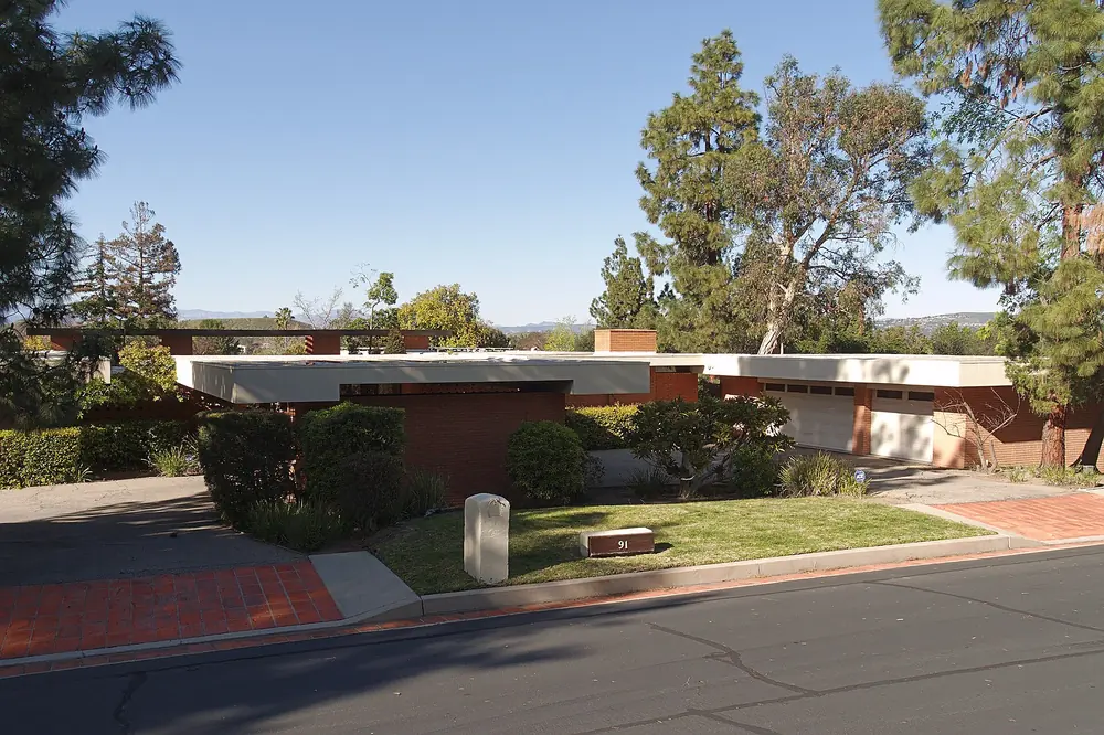 Street view of a low mid-century modern house with flat roofs behind a lawn and trees