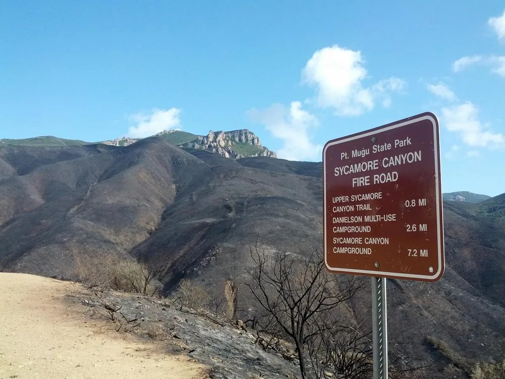 Hillside burn scar along a fire road below rocky peaks, with a park trail sign